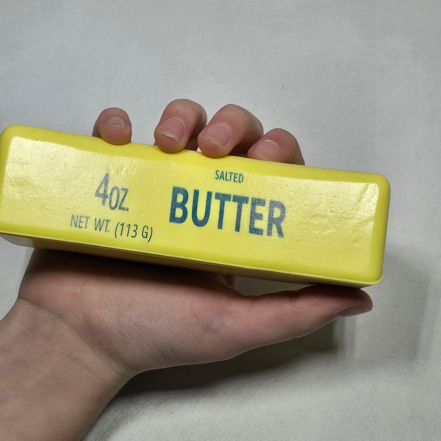A hand holding a yellow stick of salted Butter against a light gray background.