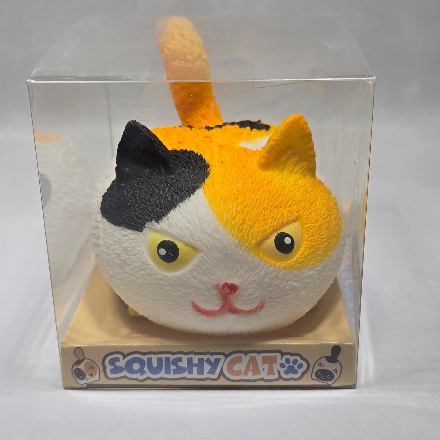 A small cat toy with yellow, white, and black patches, displayed inside a clear plastic box with a printed backing card.