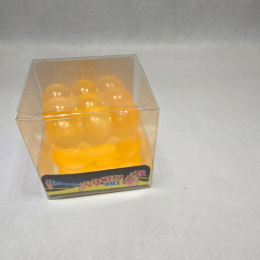A small clear plastic cube-shaped container holding several round yellow-orange gel or foam balls, photographed against a light gray textured background.
