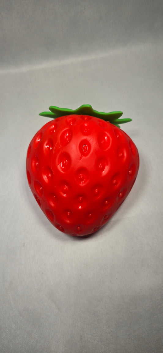 A glossy red strawberry-shaped object with green leafy tops, photographed on a plain light gray background.