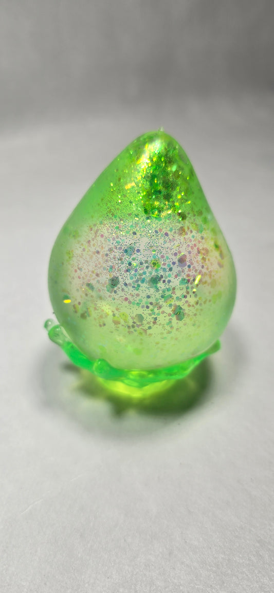 A close-up of a translucent light green cosmetic droplet or gel bead with fine glitter and iridescent flecks, resting on a light gray textured surface.