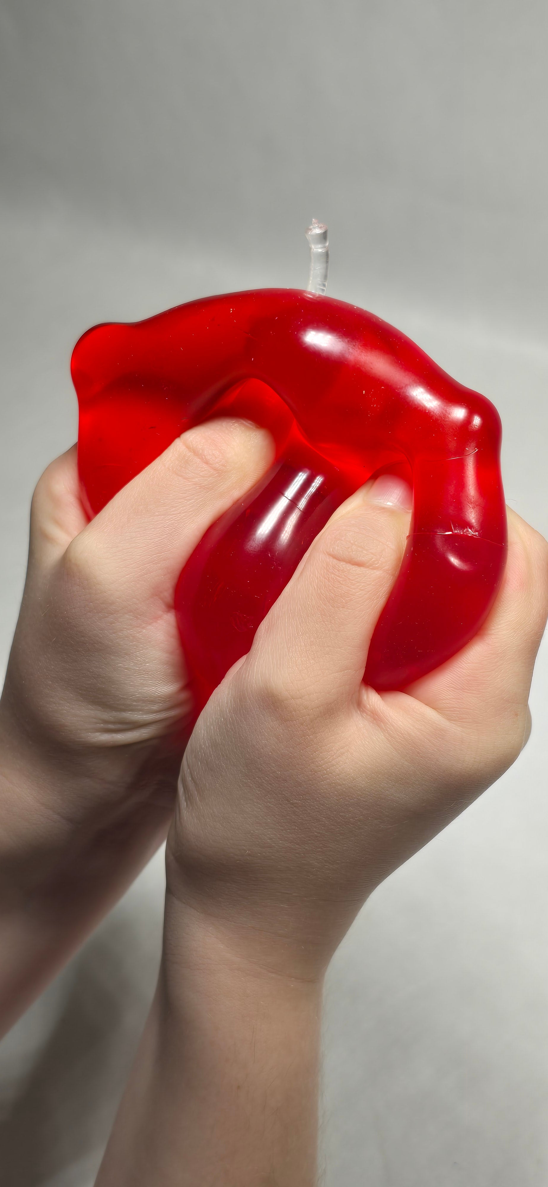 A close-up of a person's hands squeezing a translucent red, apple-shaped toy with a clear stem against a plain light gray background.