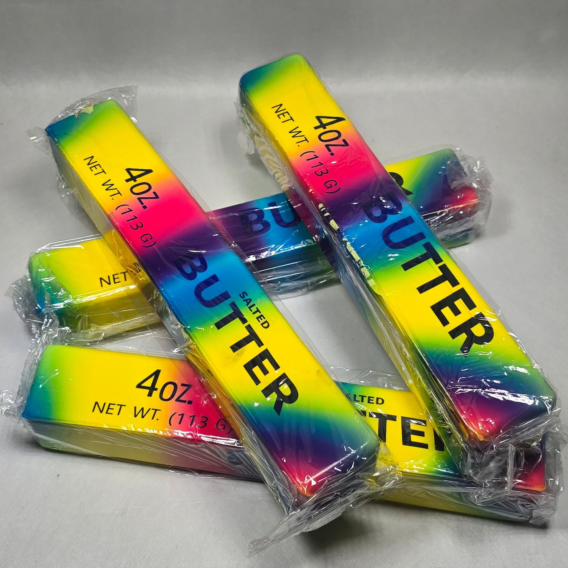 Four individually wrapped sticks of butter with a vibrant rainbow gradient label reading '4 oz' and 'BUTTER', arranged on a light gray surface.