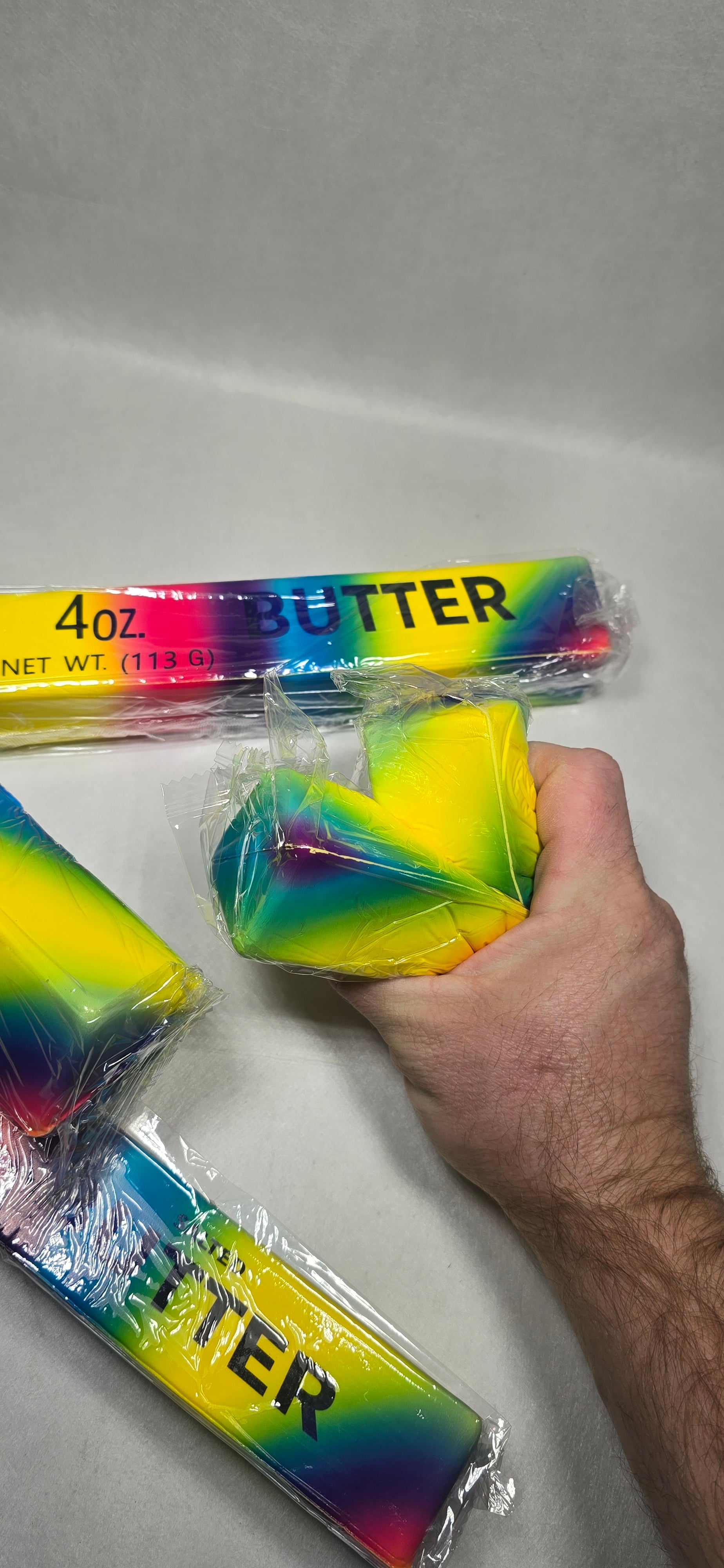 A hand holding a brightly colored, rainbow-striped rectangular item wrapped in clear plastic, with two similar packaged items on a light gray surface. The packaging shows the word "BUTTER" vertically.