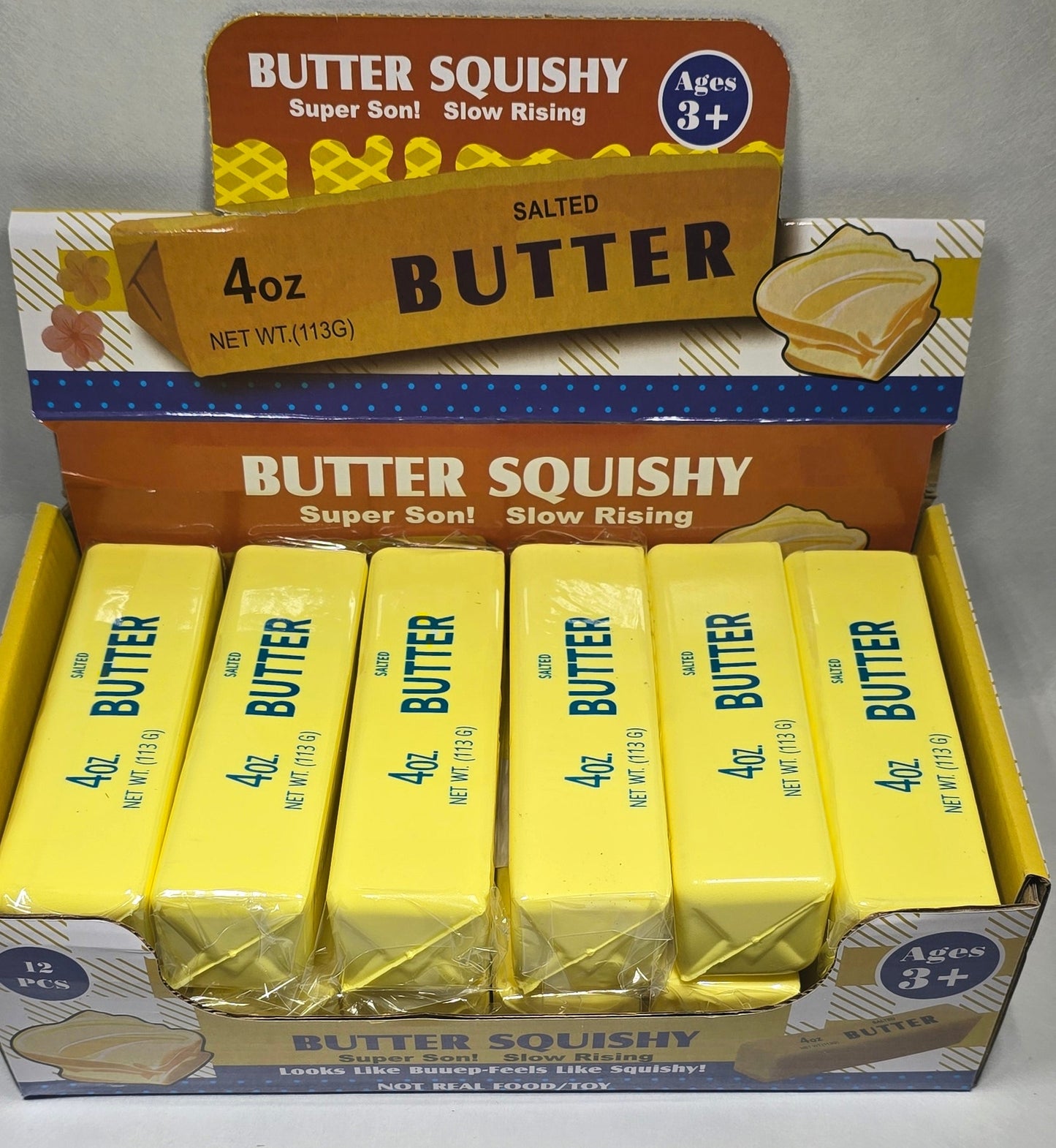 A package of Butter Squishy 4 oz butter sticks, showing several yellow butter sticks inside a box with blue and white branding, photographed on a light gray surface.