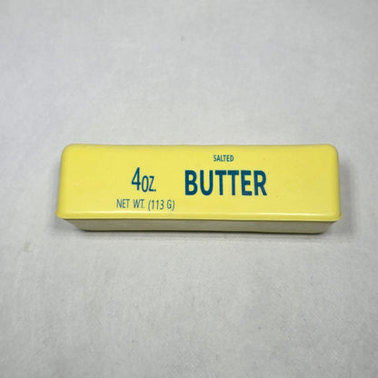 A 4 oz stick of butter in its yellow wrapper, standing vertically on a light gray textured surface.