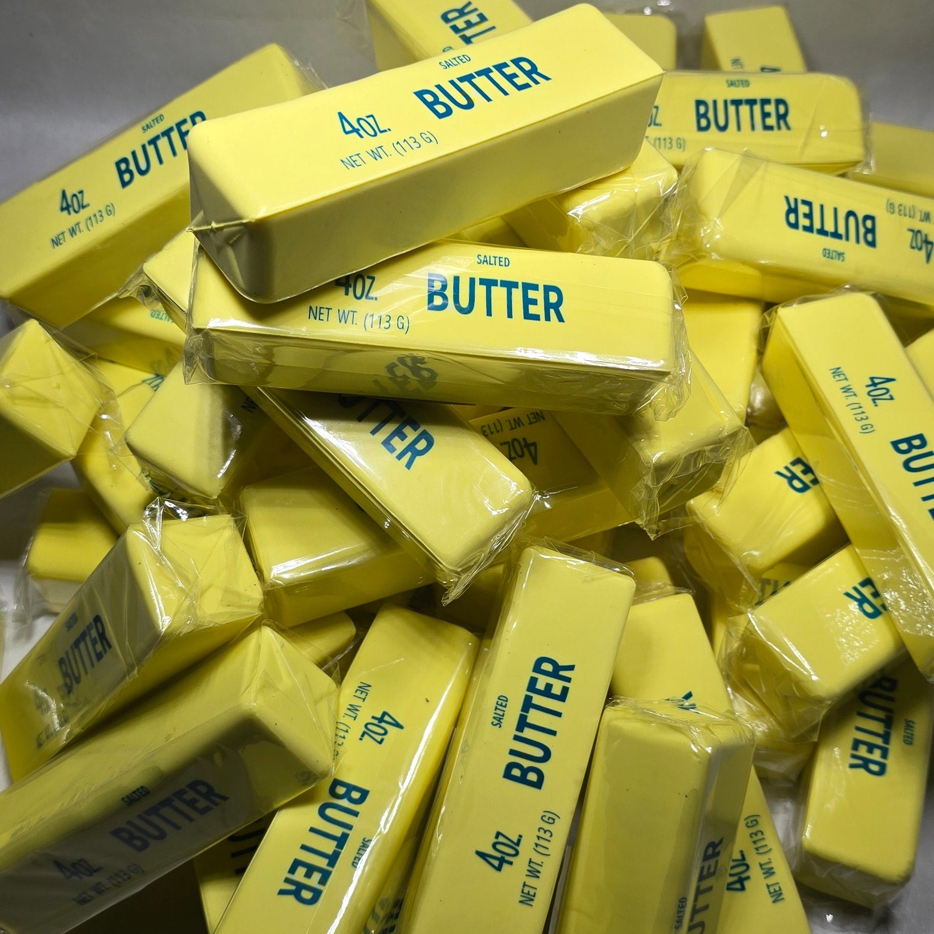 A pile of yellow butter blocks in clear plastic wrappers, with several showing the blue text 'BUTTER' on a gray background.
