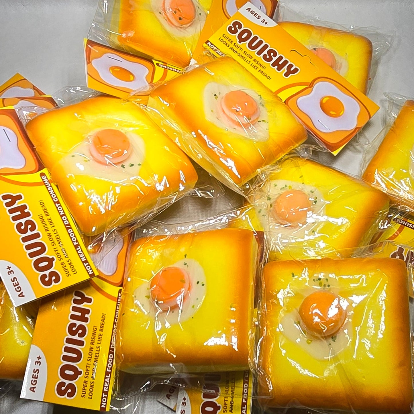 A pile of packaged novelty items shaped like toast with an egg in centers and the word 'SQUISHY' printed on them, wrapped in clear plastic on a light gray surface.