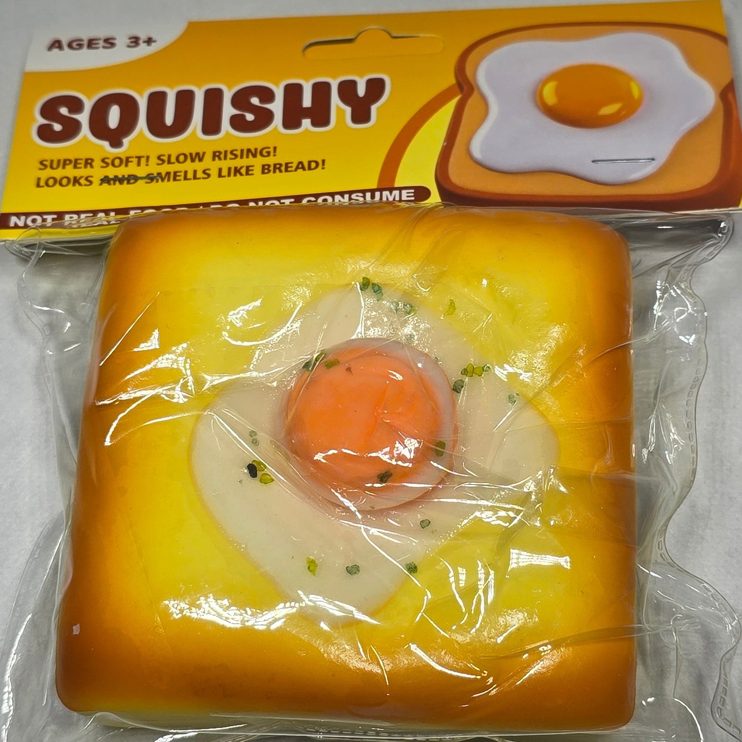 A packaged squishy toy shaped like a fried egg on a toast, still in its yellow cardboard sleeve, photographed on a light gray textured surface.