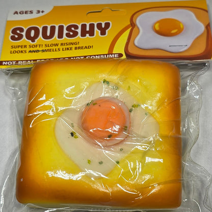 A packaged squishy toy shaped like a fried egg on a toast, still in its yellow cardboard sleeve, photographed on a light gray textured surface.