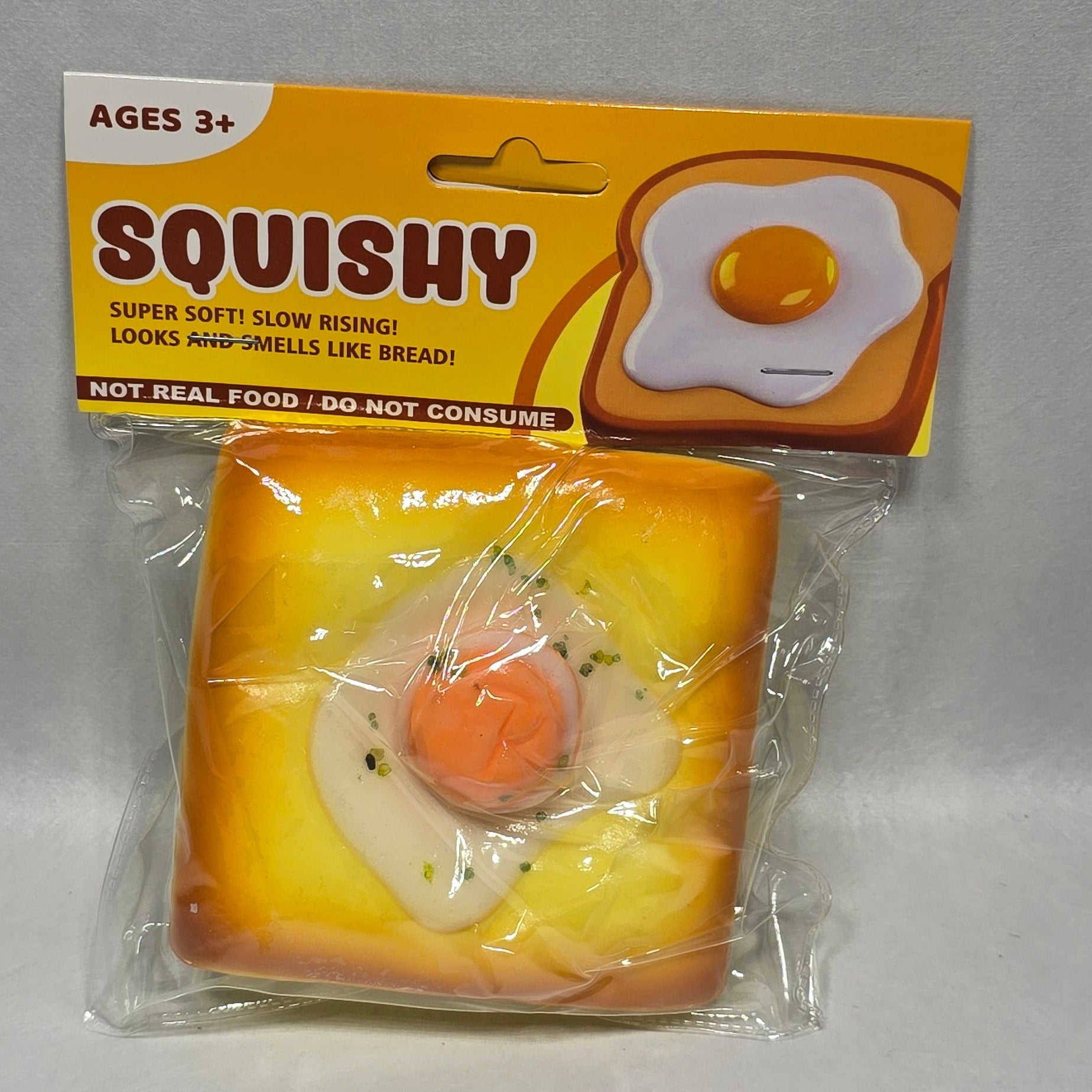 A packaged squishy toy shaped like a slice of toast with a fried egg on top, shown next to its yellow and white retail box on a light gray surface.