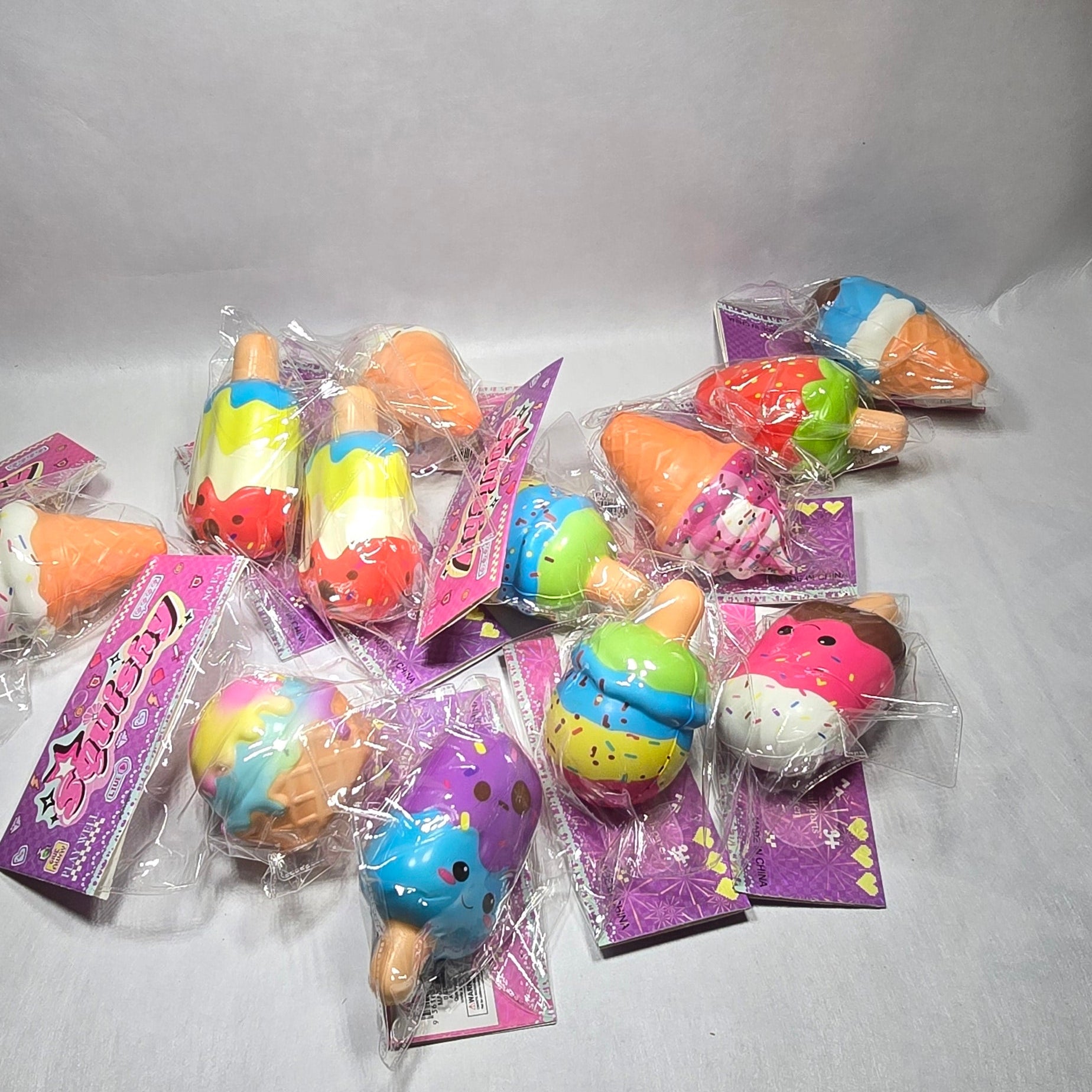 A group of small, colorful novelty squishy toys in clear plastic wrapping with purple backing cards, arranged on a light gray fabric surface.