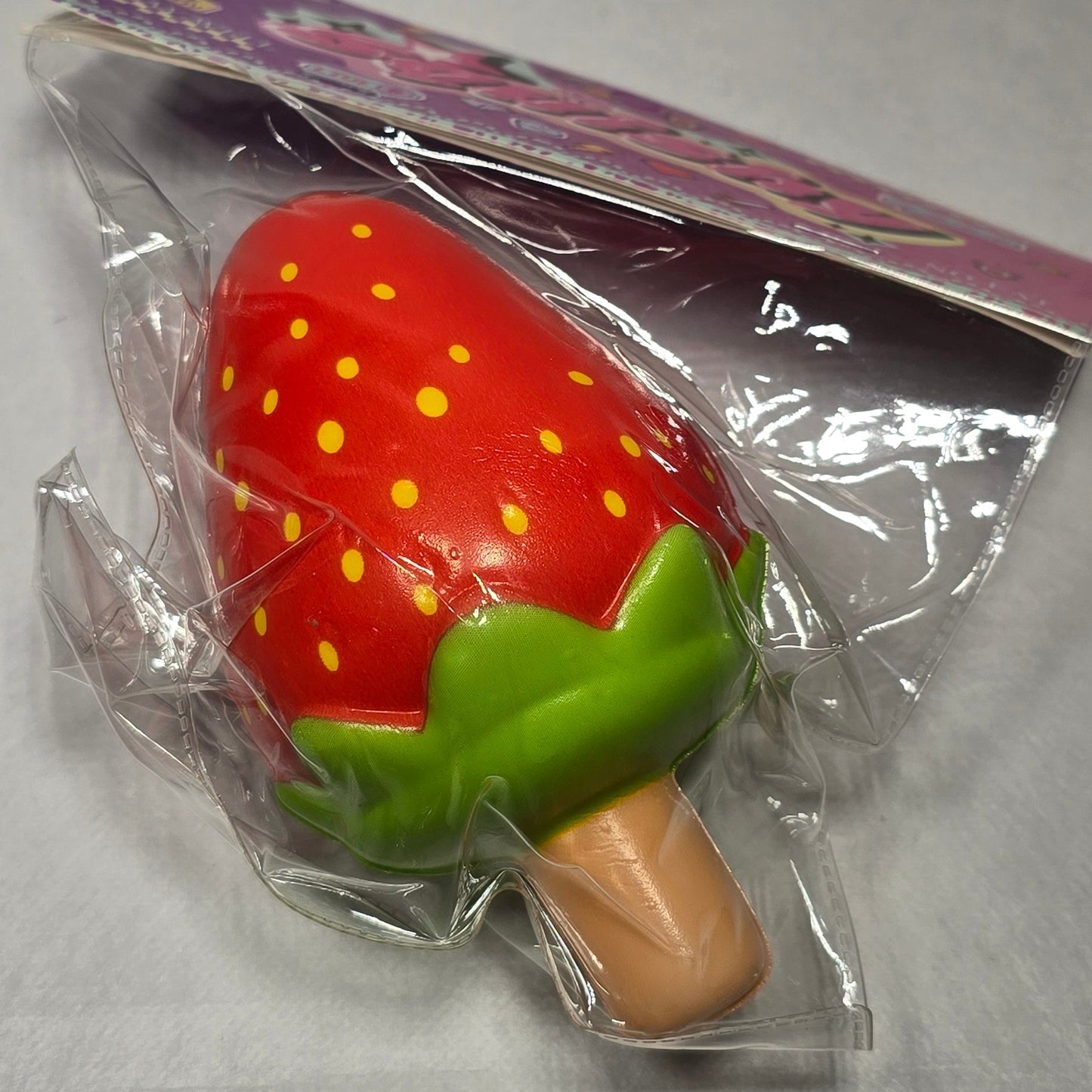 A small strawberry-shaped product wrapped in clear plastic with a purple backing card, photographed on a light gray surface.