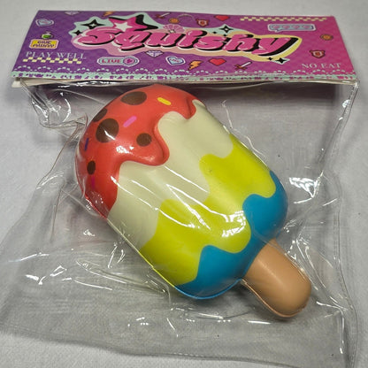A colorful novelty toy shaped like a popsicle, sealed in clear plastic packaging next to a pink and purple paper card, set against a plain light gray background.