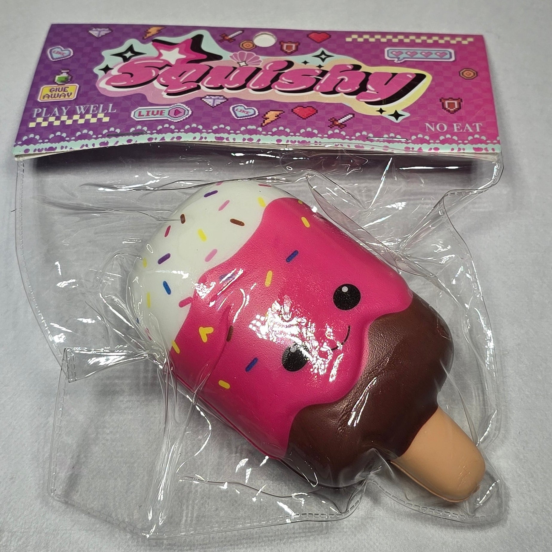 A novelty squishy toy shaped like a popsicle, packaged in clear plastic with a pink backing card. The toy has a pink body with white frosting, chocolate brown top, and a beige stick, decorated with small colorful sprinkle details.