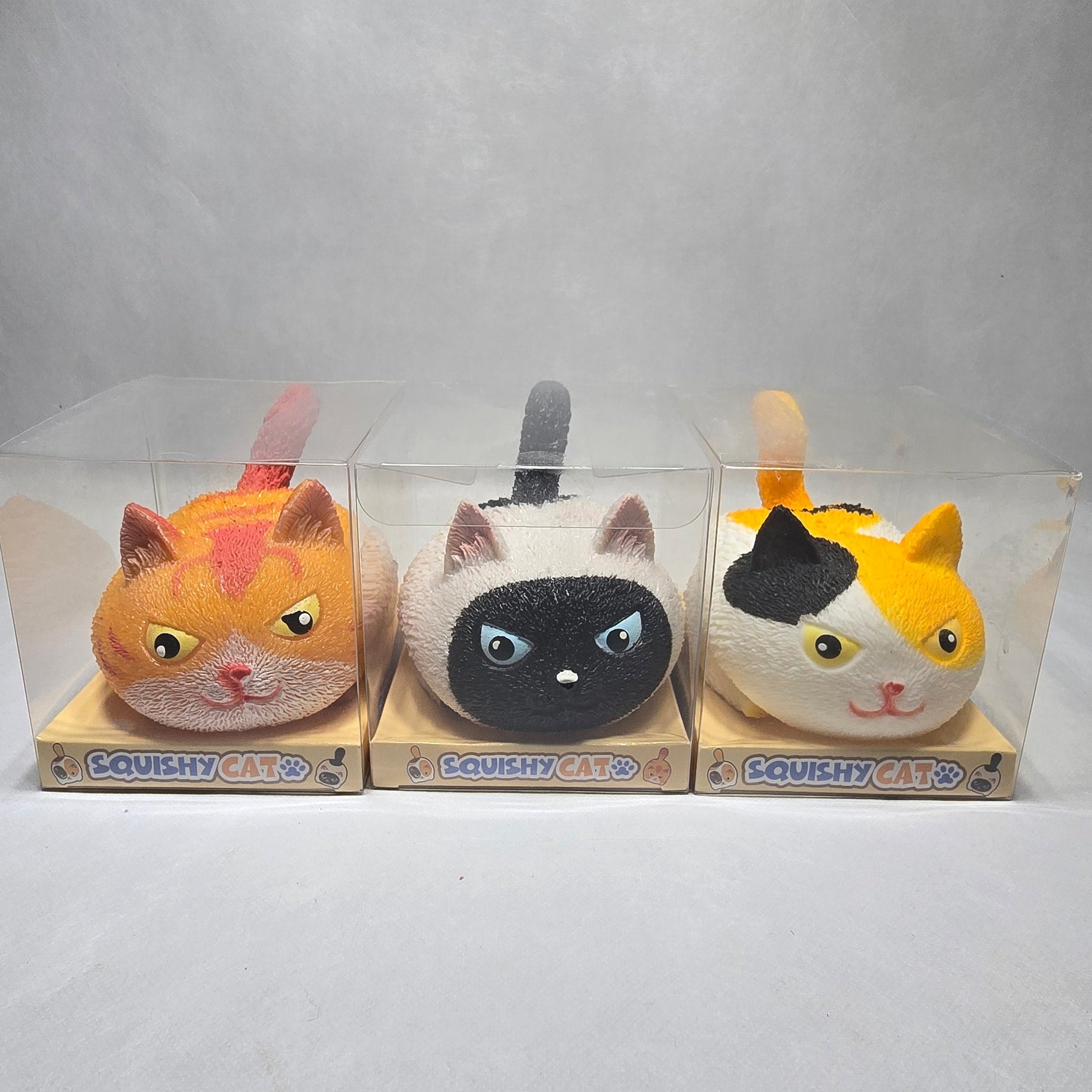 Three small cat-shaped toys displayed in a clear plastic against a light gray wall. The top figure is yellow and white with a black patch, the middle is white with a black face, and the bottom is orange with pink ears and a pink nose.