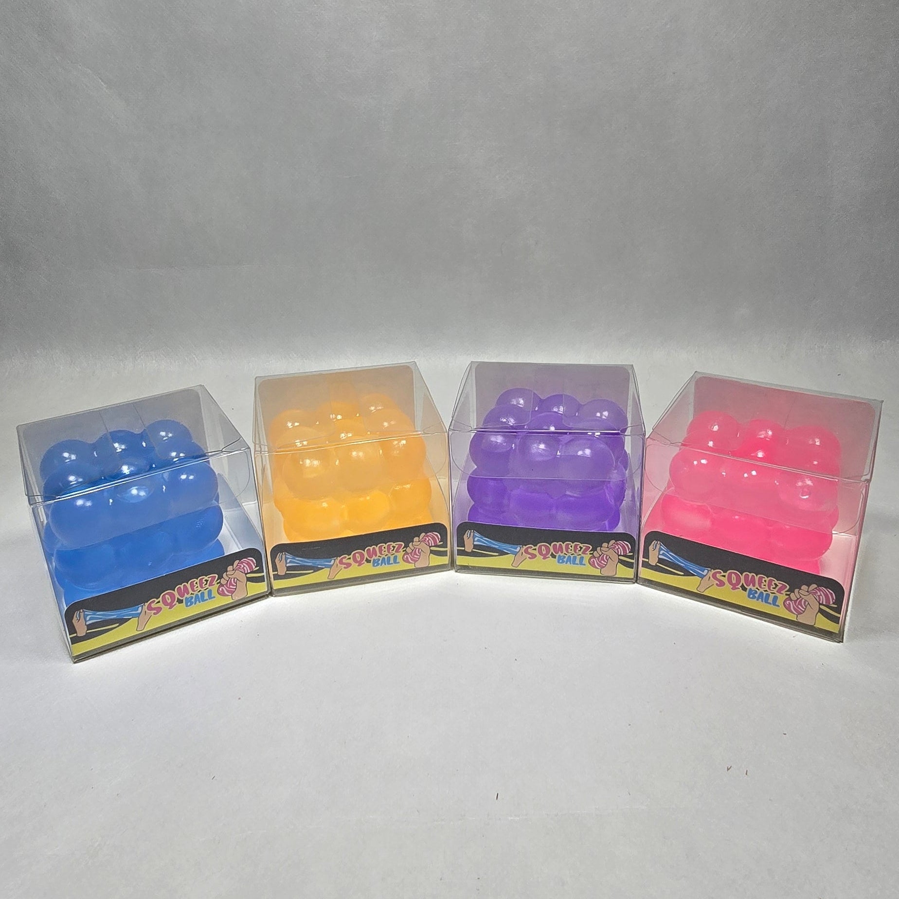 Four small clear plastic packages containing colorful rectangular items, arranged in a curved row on a light gray textured background. The packages contain pink, purple, yellow, and blue items.