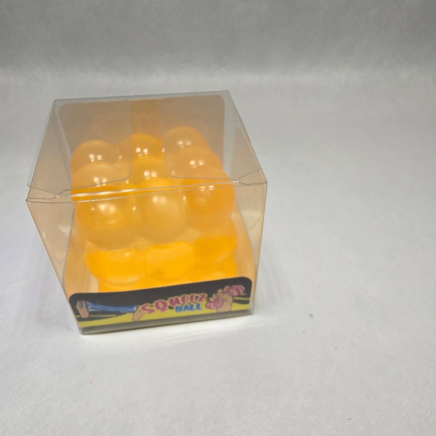 A small clear plastic cube-shaped container holding several round yellow-orange gel or foam balls, photographed against a light gray textured background.