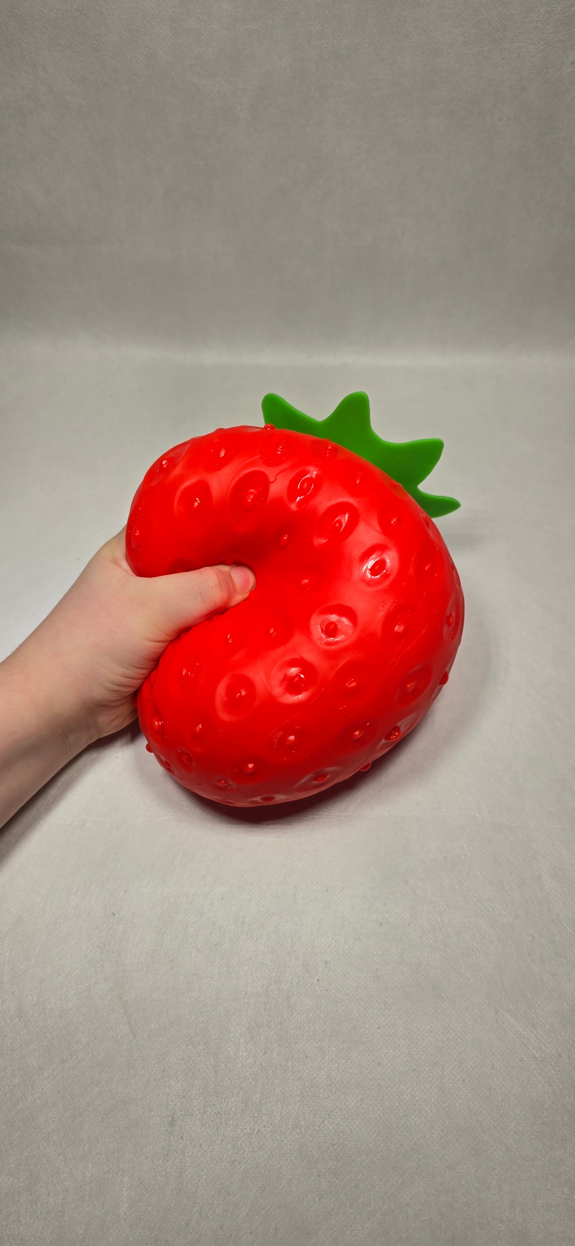 A hand holding a red strawberry-shaped squishy toy against a plain light gray background.