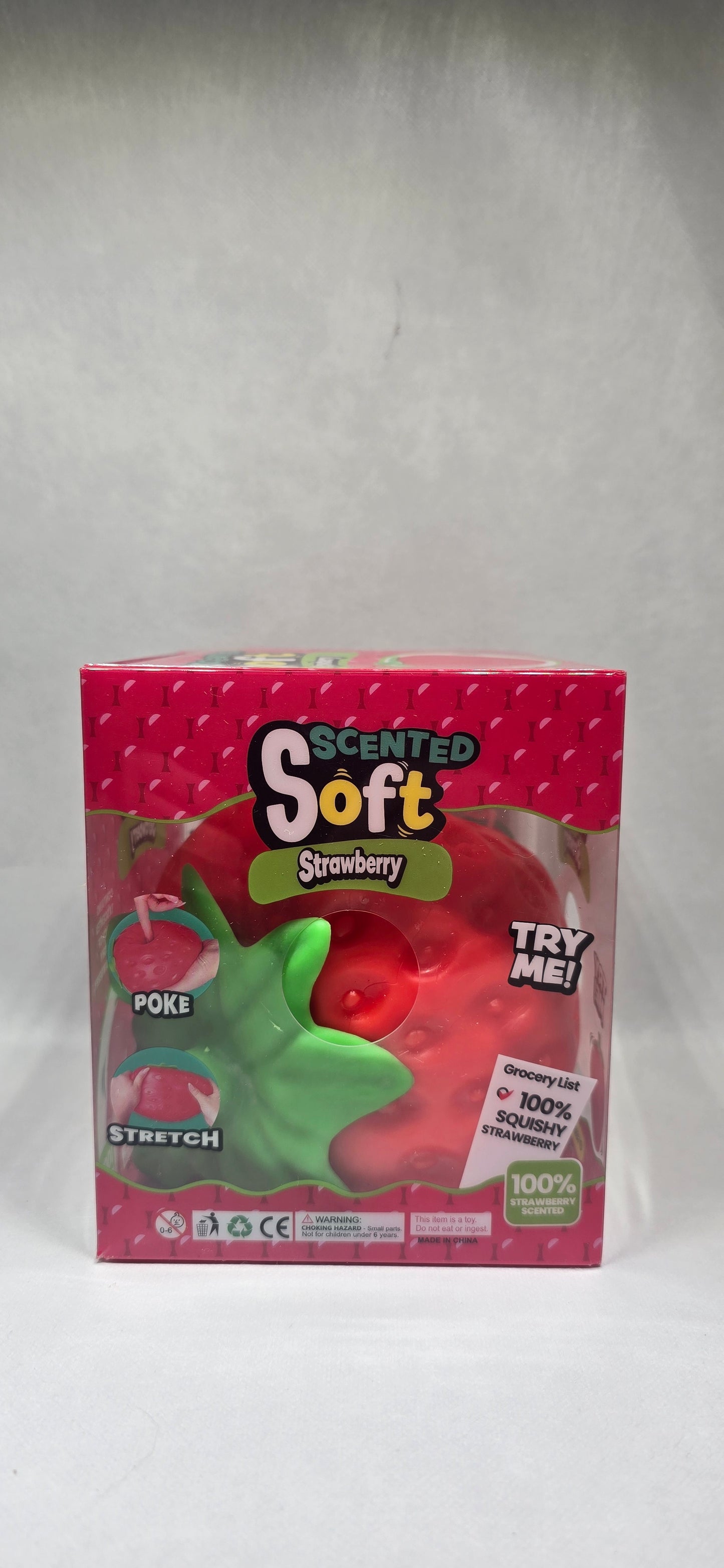 A boxed toy product labeled 'Scented Soft Strawberry' in pink packaging with a clear window showing a red strawberry-shaped item and a green leaf detail, photographed on a light gray surface.