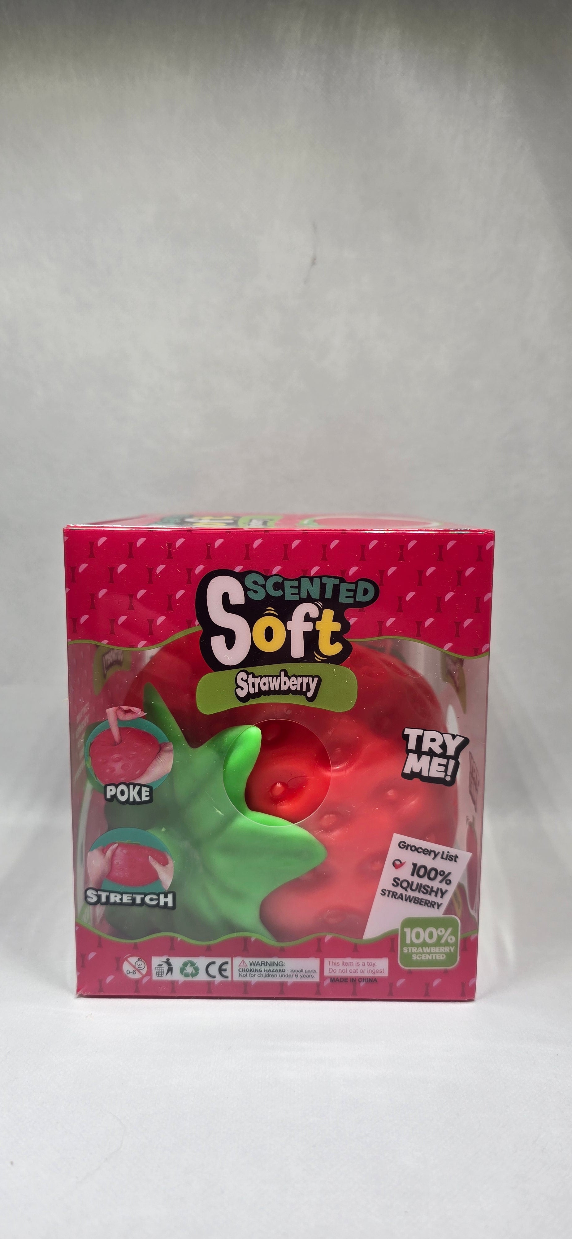 A boxed toy product labeled 'Scented Soft Strawberry' in pink packaging with a clear window showing a red strawberry-shaped item and a green leaf detail, photographed on a light gray surface.