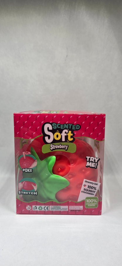 A boxed toy product labeled 'Scented Soft Strawberry' in pink packaging with a clear window showing a red strawberry-shaped item and a green leaf detail, photographed on a light gray surface.
