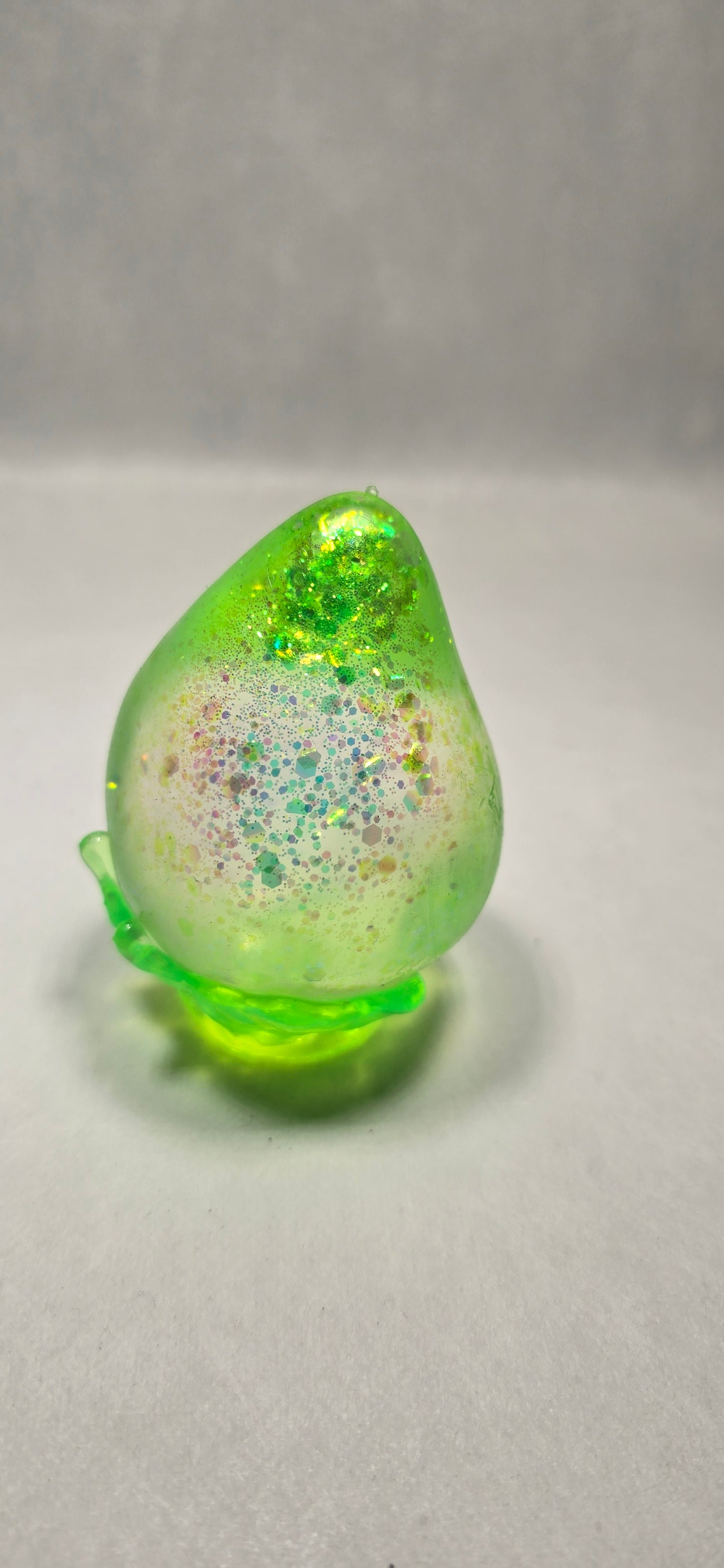 A translucent green decorative object with glittery speckles and a small attached green stem, photographed against a plain light background.