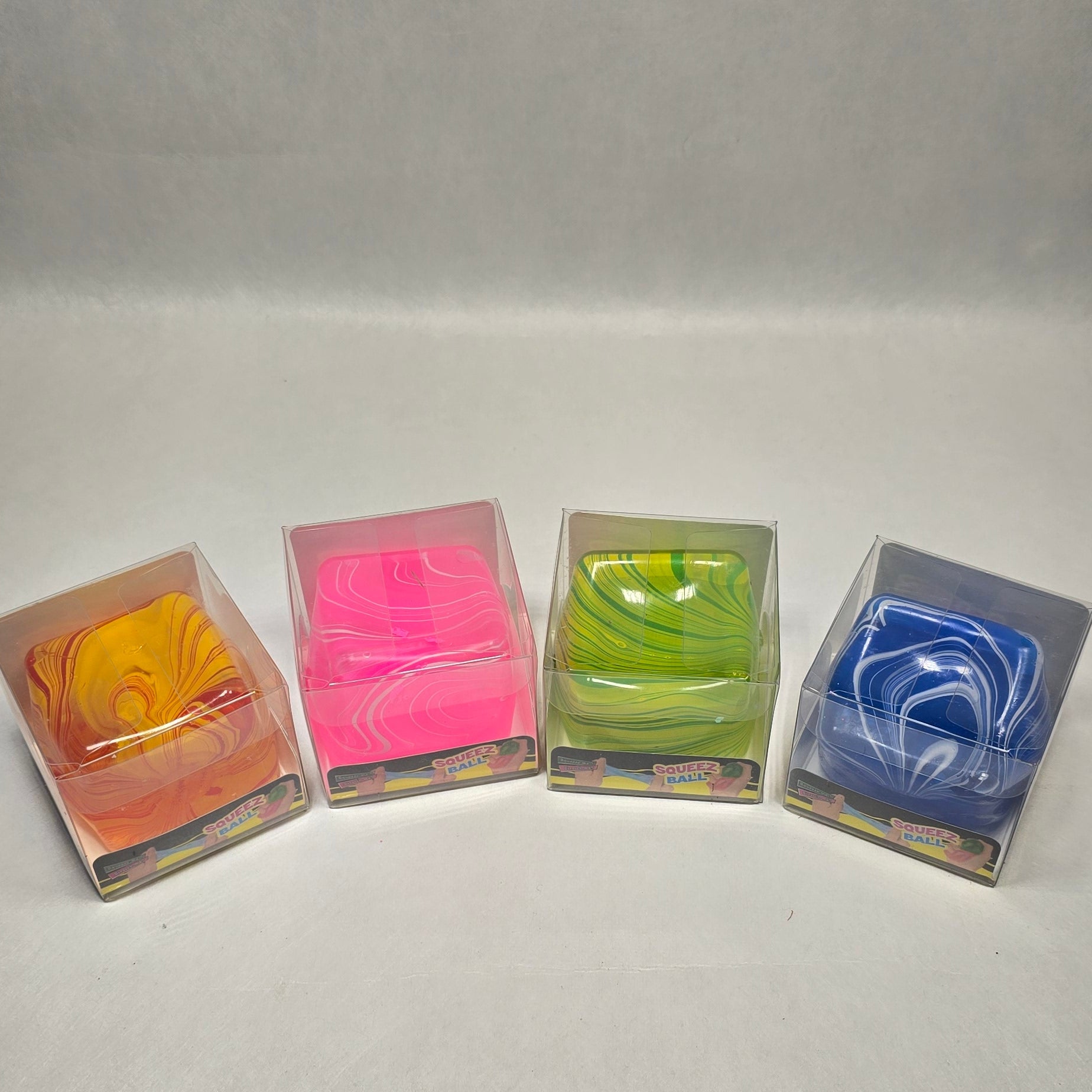 Four small square plastic packages containing colorful folded items, arranged in a row on a light gray fabric background. The packages contain orange, pink, green, and blue items.