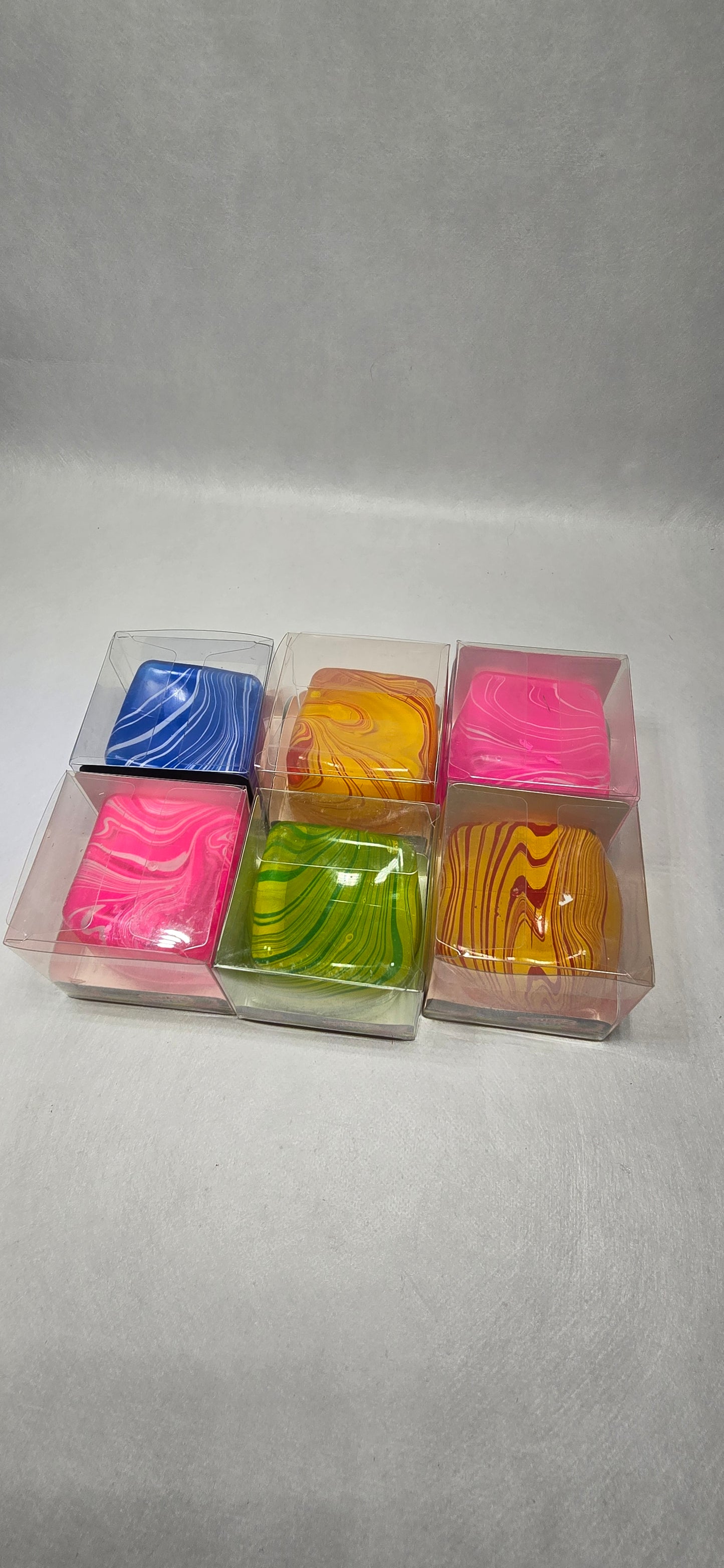 Six small square clear plastic boxes containing colorful marbled soap bars, arranged in two rows on a light gray textured background.