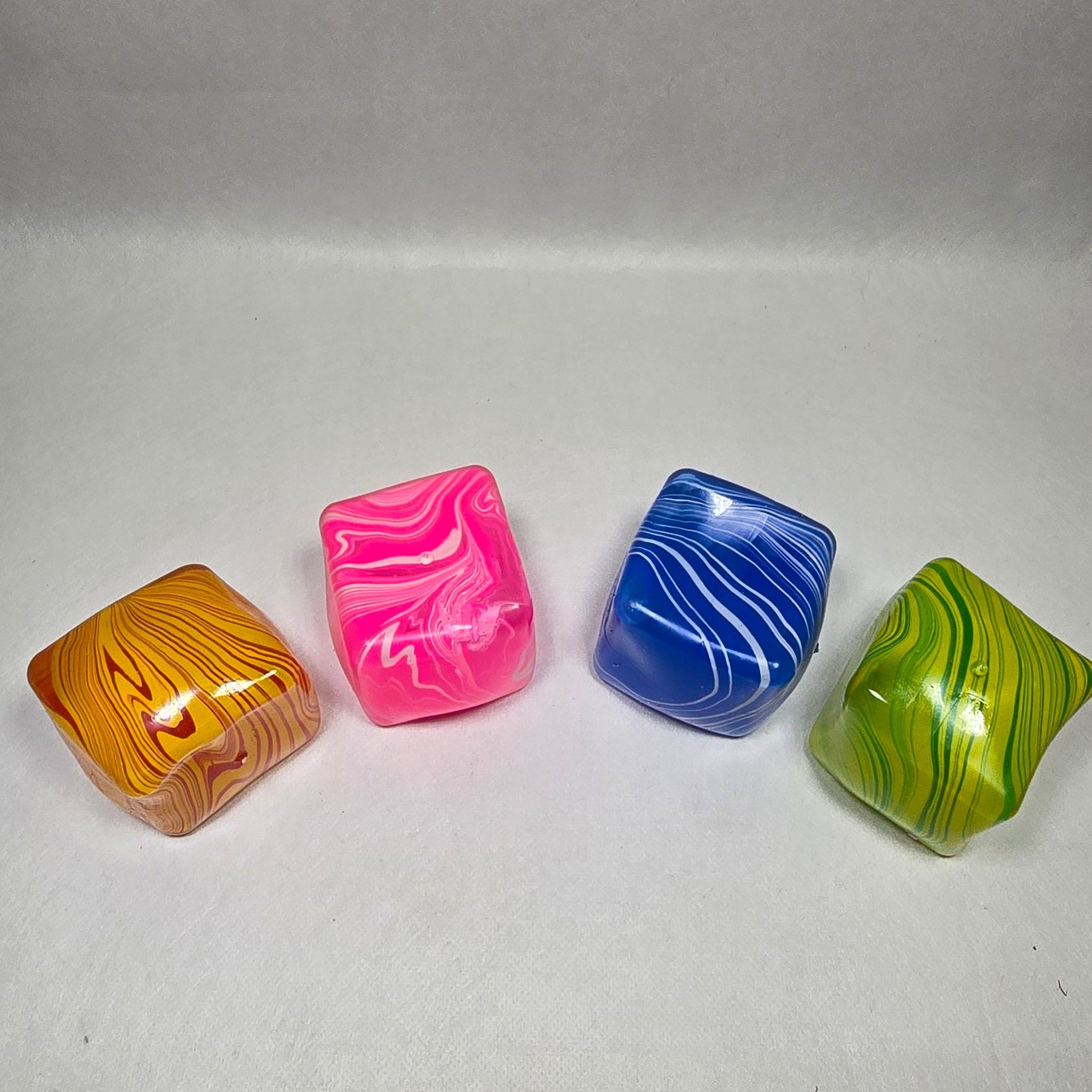 Four small cubes with rounded corners, featuring marbled swirls of orange, pink, blue, and green, arranged in a row on a light gray background.