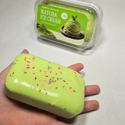 A hand holding a lime green squishy slime bar with multicolored sprinkles next to its clear plastic packaging container on a white surface.