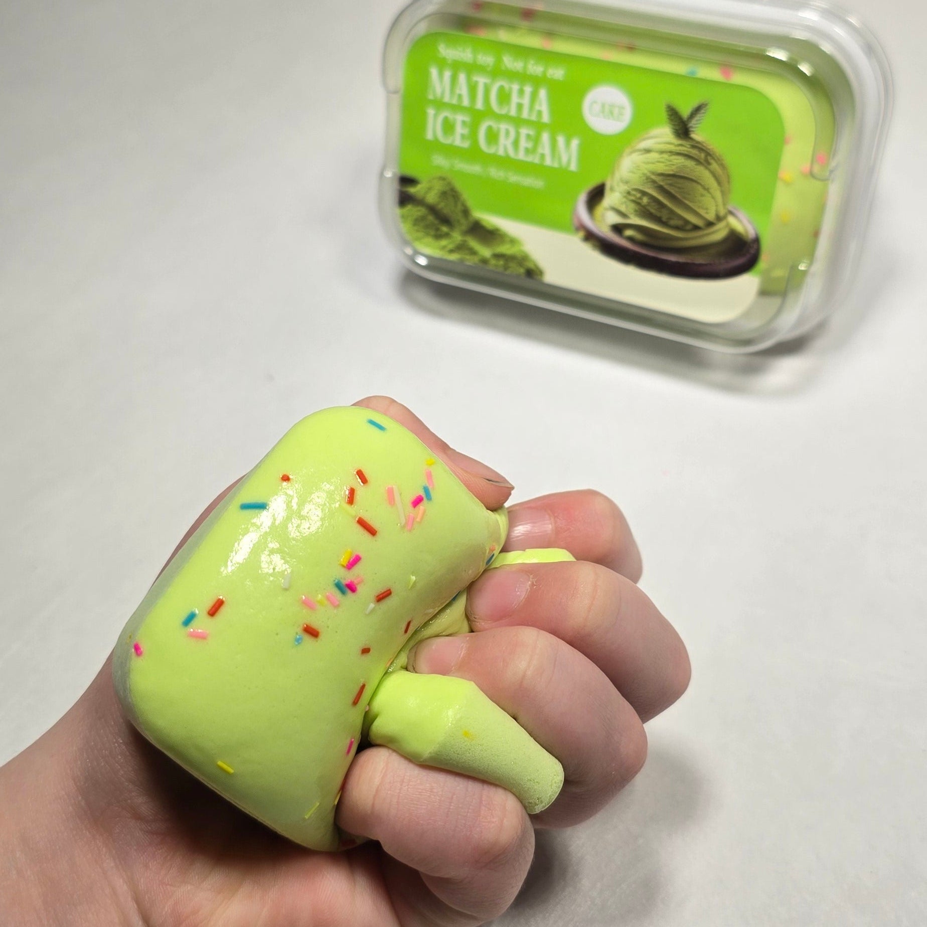 A hand squeezing a light green squishy toy with colorful sprinkles, next to its clear plastic packaging container on a light gray textured background.