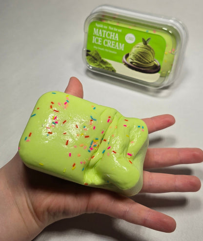 A hand holds a bright yellow-green squishy slime bar with colorful sprinkles, next to its clear plastic packaging container on a light gray surface.