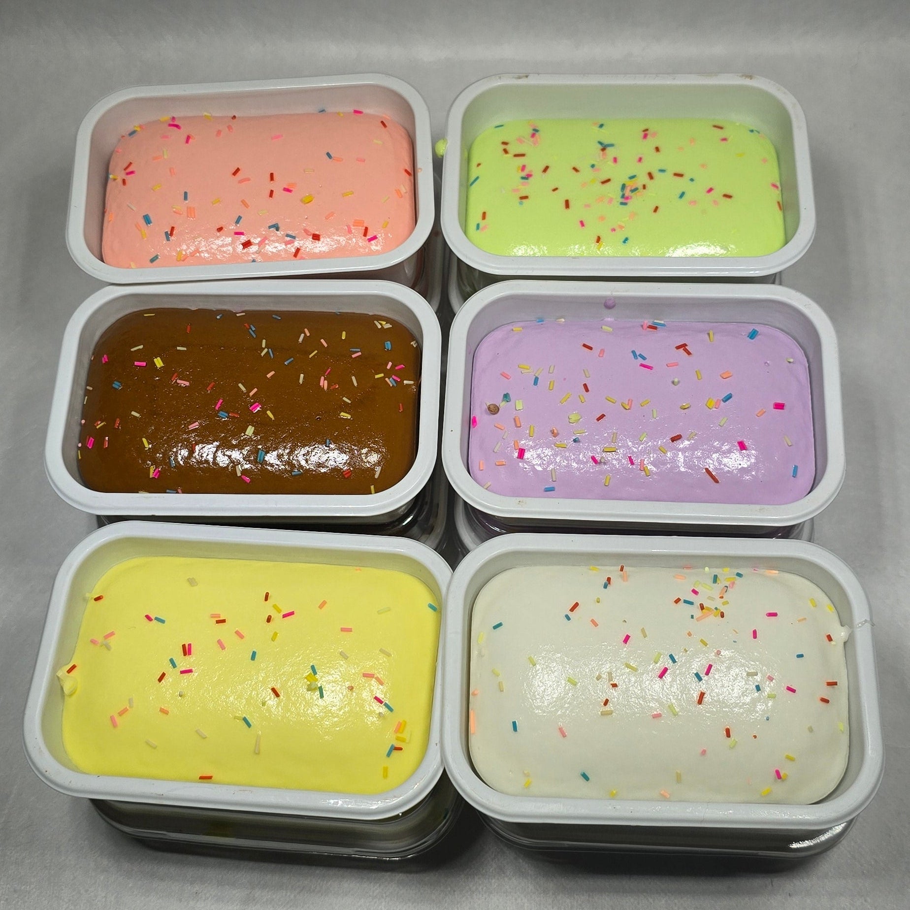 Six small rectangular plastic containers filled with different colored gel or cream products, arranged in two rows on a gray surface. The colors include light green, lavender, off-white, peach, dark brown, and pale yellow, with some sprinkles visible on several containers.