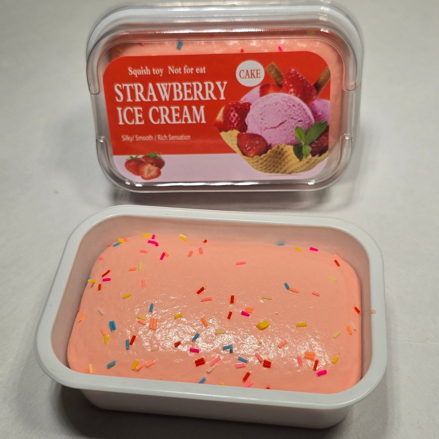 Two small plastic containers of strawberry ice cream on a light gray surface, one showing the red-labeled product packaging and the other showing the pale pink ice cream with colorful sprinkles.