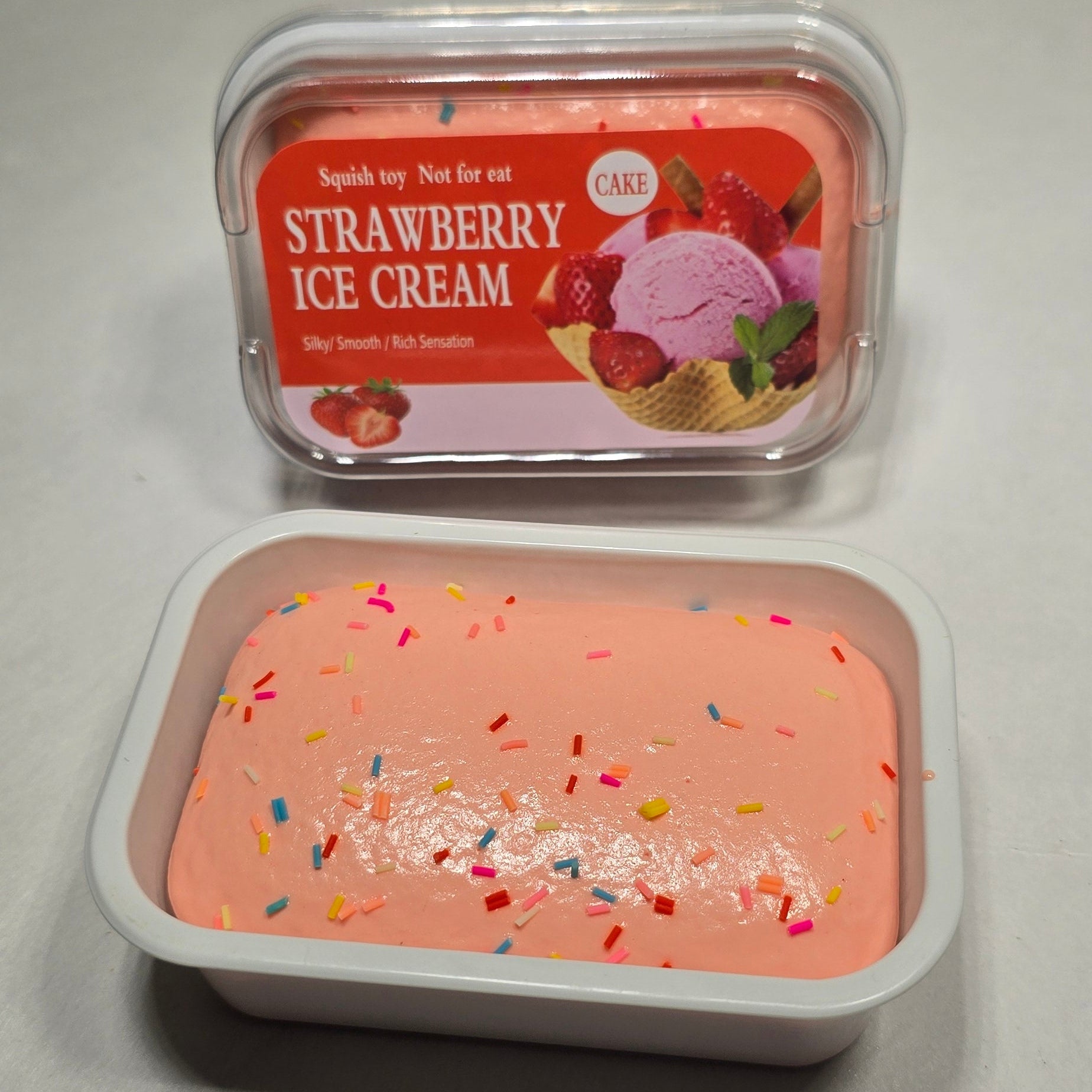 Two small plastic containers of strawberry ice cream on a light gray surface, one showing the red-labeled product packaging and the other showing the pale pink ice cream with colorful sprinkles.